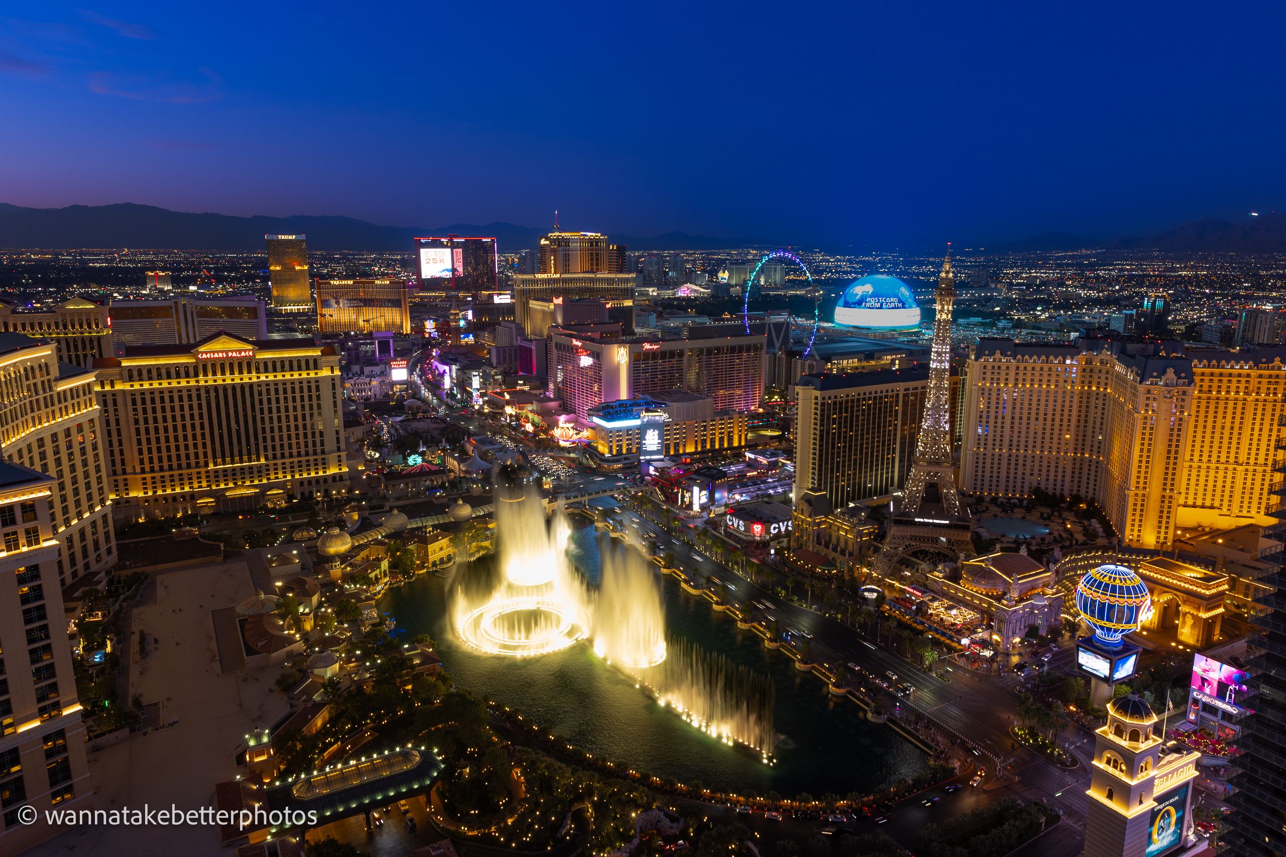 Elevated view of the Las Vegas Strip beneath a deep blue sky, with illuminated Bellagio fountains in the foreground and glowing hotels and the Sphere beyond