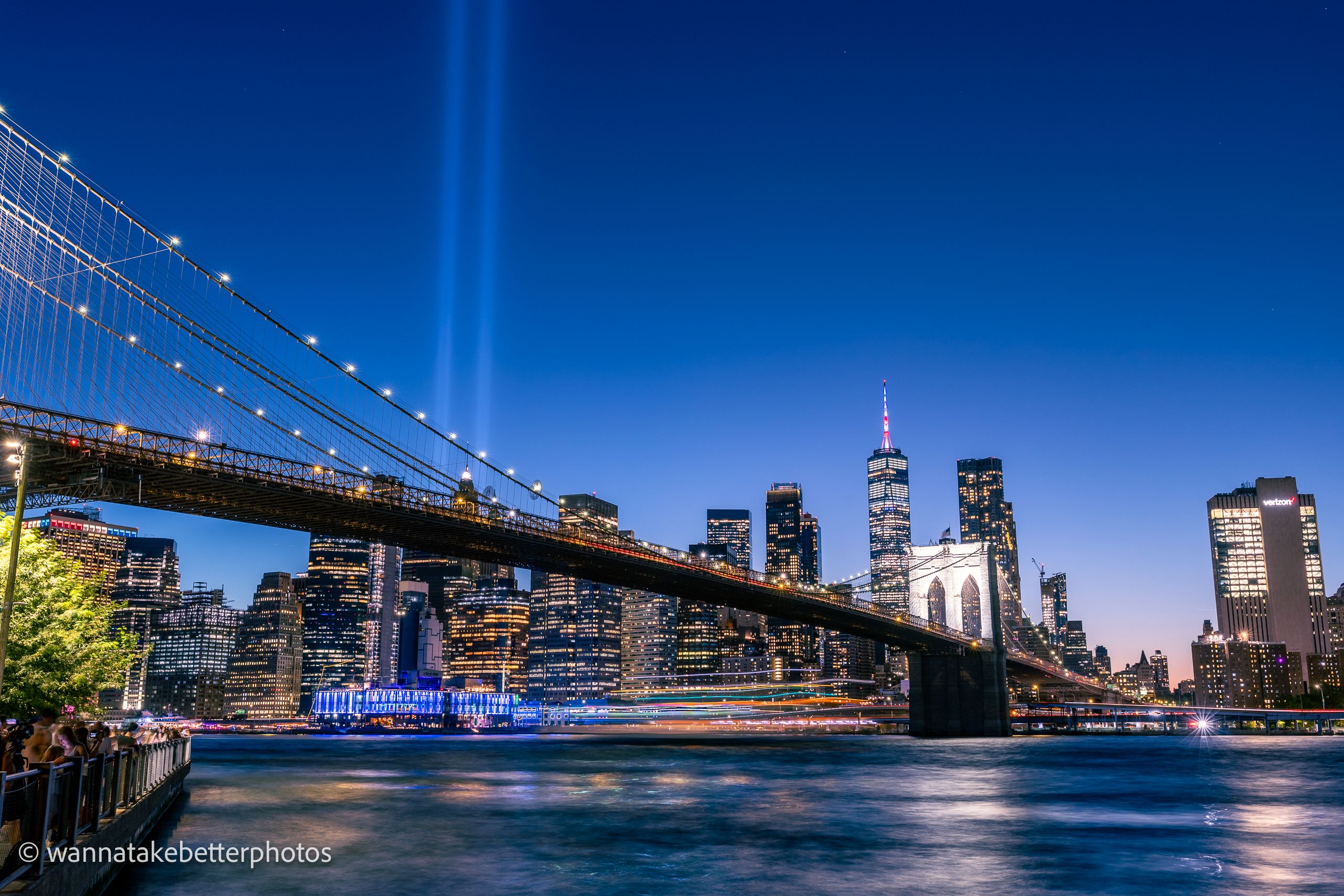 Brooklyn Bridge crossing the East River toward Lower Manhattan at blue hour, with the twin Tribute in Light beams above the skyline and boat light trails across the water